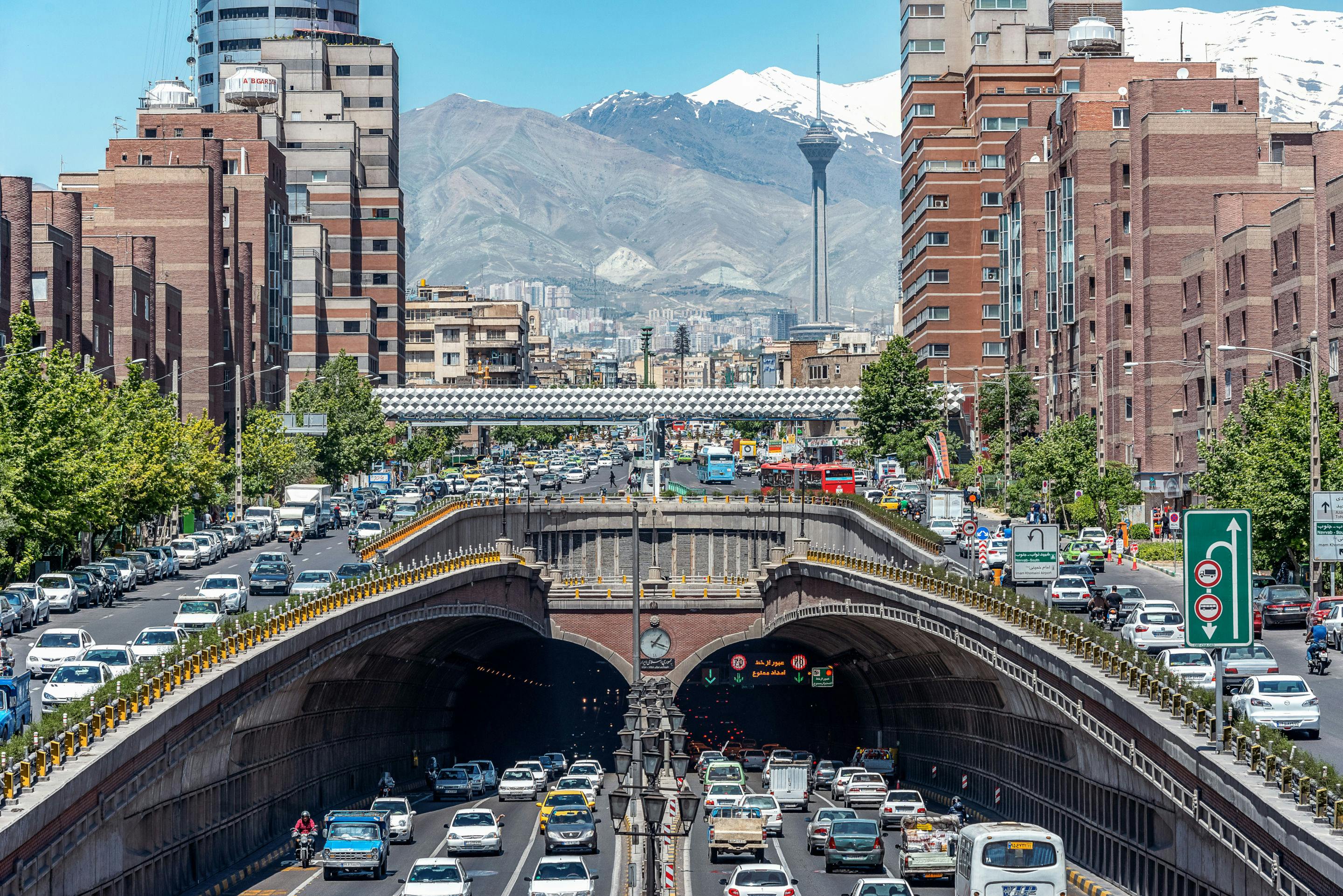 Traffic in Tehran, Iran looks like a dozen lanes each way across a soaring bridge and tunnel system, with motorcyles, international tourists, businessmen, autobuses, and even pedestrians. The image is framed against a mountainous backdrop  found in these areas of the Middle East and Central / West Asia.